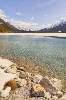 Portrait view across goat pond in winter with rocks in the foreground and Rockie Mountains in the background, Kananaskis, Alberta clipart