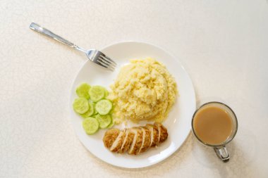Couscous on a white plate with sliced cucumber and cutlet, next to coffee with milk. 
