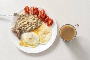 Breakfast of oatmeal with roasted egg, tomato slices, herring slices and a cup of coffee with milk. 