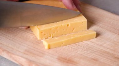 Knife in mans hand cuts yellow cheese with holes in cubes and slices. 