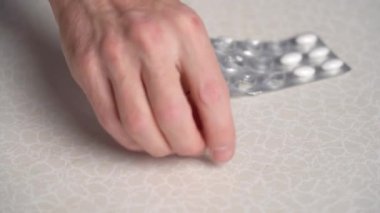 Mans hand takes white tablet lying near blister from table and points to camera. 