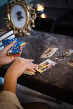 Elektrikli mavi oje ve metalik mücevherlerle süslenmiş bir kadın masanda şık bir bilek aksesuarıyla ustaca tarot kartları oynuyor.