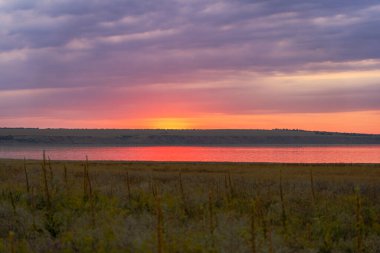 Canlı günbatımı gökyüzü, mor ve turuncu renkler ile bir nehre yansıyor, çimenli bir çayırdan görünüyor, tepeler ve dramatik bulutlar yukarıda. 