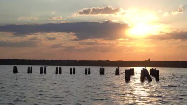 Gün batımında nehrin akşam manzarası, arka planda altın güneş ışığı, sakin su ve bulutları yansıtan kırık ahşap direkler. 