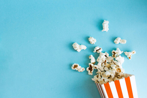 Red and white stripped popcorn box with a popcorn around on a blue background. TV watching concept with copy space. Movie night. Entertainment concept.