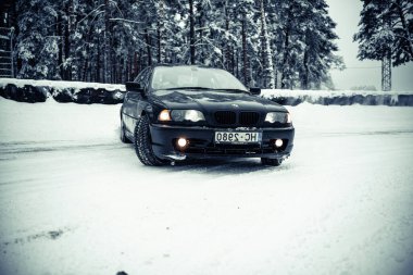 12-12-2022 Riga, Letonya. Karlı bir yola park etmiş siyah bir araba. Ağaçların ve çalıların önünde.. .