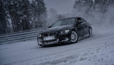 12-12-2022 Riga, Letonya. Karla kaplı bir yolda ilerleyen siyah bir araba. Arka planda ağaçlar ve ön planda bir çit var.. .