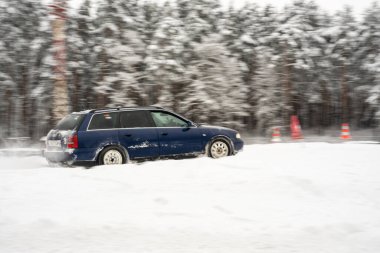 12-12-2022 Riga, Letonya. Arka planda ağaçlar ve kırmızı bir trafik konisi olan karla kaplı bir orman bölgesinden geçen mavi bir araba.. .