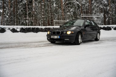 12-12-2022 Riga, Letonya. Karlı bir yolda ilerleyen siyah bir araba. Ağaçlardan oluşan bir ormanın yanında.. .