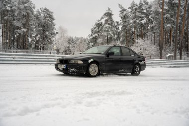 12-12-2022 Riga, Letonya. Karla kaplı bir yolda ilerleyen siyah bir araba. Ağaçlardan oluşan bir ormanın yanında ve üzerinde kar olan bir çit.. .