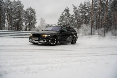 12-12-2022 Riga, Letonya. Karla kaplı bir yolda ilerleyen siyah bir araba. Ağaçların yanında ve üzerinde kar olan bir çit.. .