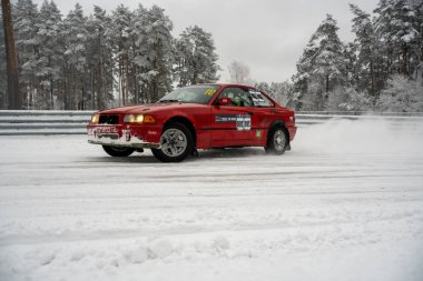 12-12-2022 Riga, Letonya. Karla kaplı bir yolda ilerleyen kırmızı bir araba. Ağaçlardan oluşan bir ormanın yanında ve üzerinde kar olan bir çit.. .