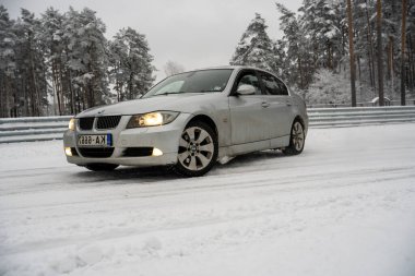 12-12-2022 Riga, Letonya. Karla kaplı bir yolda ilerleyen gümüş bir araba. Ağaçlardan oluşan bir ormanın yanında ve üzerinde kar olan bir çit.. .