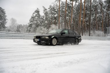 12-12-2022 Riga, Letonya. Karla kaplı bir yolda ilerleyen siyah bir araba. Ağaçlardan oluşan bir ormanın yanında ve üzerinde kar olan bir çit.. .