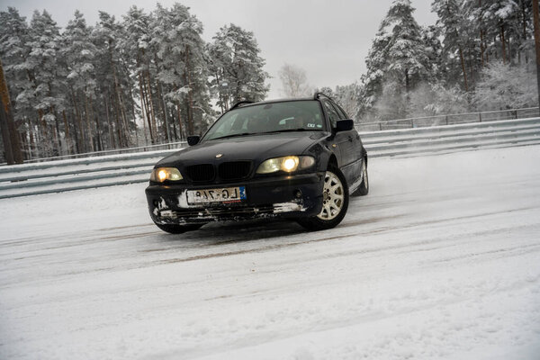 12-12-2022 Riga, Latvia a black car driving down a snowy road next to a forest of trees and a fence with snow on it. .