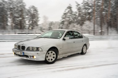 12-12-2022 Riga, Letonya Karlı bir yolda ilerleyen beyaz bir araba Ağaçların yanında ve kenarları karlı bir çit. .