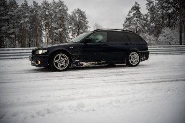 12-12-2022 Riga, Letonya. Çitin önünde karlı bir yola park etmiş siyah bir araba ve arka planda karlı bir yer olan ağaçlar.. .