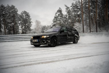 12-12-2022 Riga, Letonya. Karla kaplı bir yolda ilerleyen siyah bir araba. Ağaçların yanında, üzerinde kar olan bir çit ve yol kenarına park etmiş birkaç araba.. .