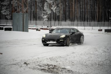 12-12-2022 Riga, Letonya. Arka planda ağaçlar ve ön planda çitlerle kaplı, karla kaplı bir yolda ilerleyen siyah bir araba.. .