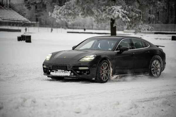 12-12-2022 Riga, Latvia a black porsche panamer driving through the snow in a park area with a bench and trees in the background. .