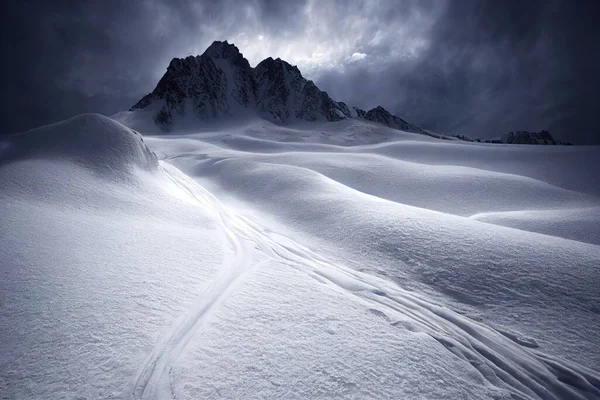 a snow covered mountain with a trail going through it\'s snow covered ground and a cloudy sky above. .
