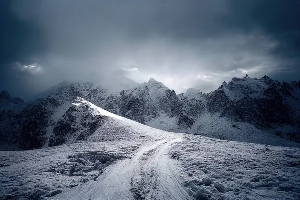 a snow covered mountain with a trail going through it and a cloudy sky above it with sun shining through the clouds. .