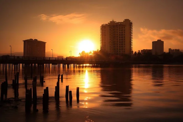 the sun is setting over the water near a city with tall buildings and a pier in the foreground, with the sun reflecting off the water in the distance.