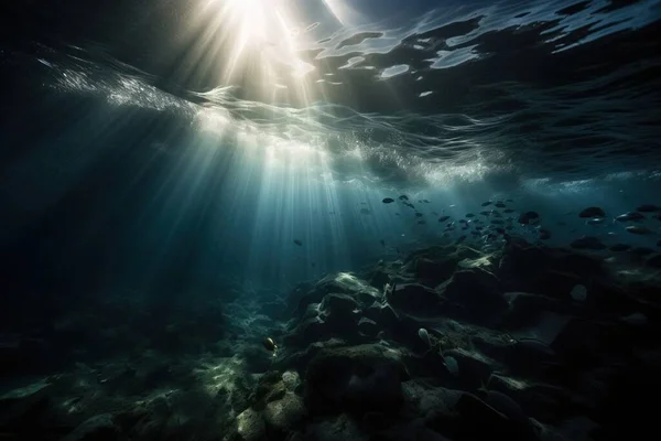 sunlight shining through the water onto a rocky reef with fish swimming below it and a rock wall below the water surface with rocks and pebbles.