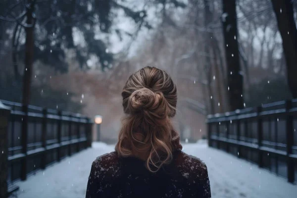 a woman standing on a bridge in a snowy park with her hair in a bun in the wind and snow falling on the ground behind her.