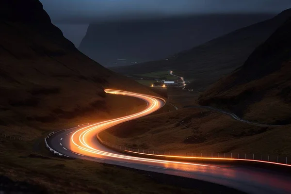 a long exposure photo of a road in the mountains at night with a car light streaking down the road and a house in the distance.