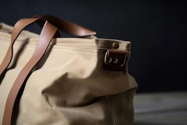 a tan canvas bag with a brown leather handle and a brown leather strap on the side of the bag, with a dark background of wood.