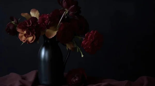 a black vase filled with red flowers on top of a bed of red sheets on a black background with a black background behind it and a dark background.
