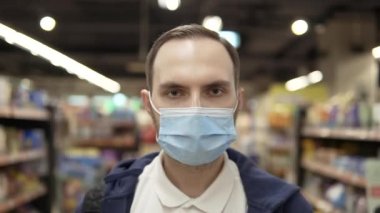 Portrait of a young man in a protective medical mask in a supermarket. He looks at the camera. Close-up. Pandemic COVID-19.