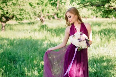 A bridesmaid in a green summer garden poses in a purple dress with a bouquet of light peonies and holds the hem of the dress with her hand. Copy space.