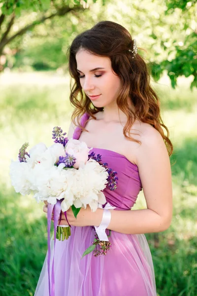 Young woman with professional makeup and hairstyle in a purple dress holds a bouquet of white peonies with her eyes closed in a green garden in the summer. High quality shot