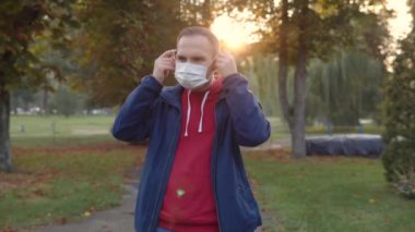 A young man in a red hoodie and a blue windbreaker wears a protective medical disposable mask on his face when entering a public place. Anti-Covid restrictions in the autumn park. Ultra HD video.