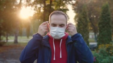 A man in a public place in the park puts a disposable protective medical mask on his face and looks into the camera. Anti-covid measures during lockdown. 4k footage. 