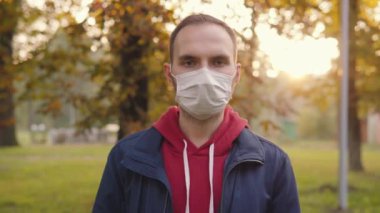 Portrait of a young man in a medical disposable protective mask. A man stands in an autumn public place in a park and looks at the camera. Anti-covid measures.