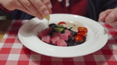 A man squeezes lemon juice on raw tuna. Italian cuisine: black pasta with cuttlefish ink, fresh tuna, cherry tomatoes, fish sauce. Close-up view.