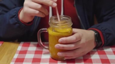 Man in a red hoodie and blue jacket stirs vitamin sea buckthorn tea, drinks a hot warming drink through a plastic straw. Traditional medicine concept. Close-up shot.