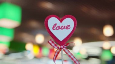 A concept Valentines day. Decor, toy, heart-shaped attribute on a stick with a bow and the word love against the multi-colored bokeh, blurred background of the restaurant. Close-up shot, 4k footage.