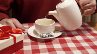 Male hand pours black or green tea from a white porcelain teapot into a white cup on the table, gift box with red bow on the left side of the frame. High quality 4k footage. 