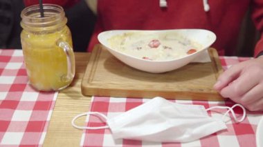  Vaccinated man eats turkey julienne at a restaurant during the pandemic. Anti-covid measures, disposable medical mask lies on the table next to the sea buckthorn vitamin drink. Close-up view.