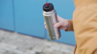 Side view of a male tourist slowly drinks a hot drink from the lid of a steel thermos. Close up shot of mans hands.