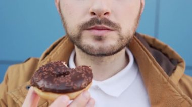 Close-up, a man with a mustache and a beard eats a chocolate doughnut against a blue background.