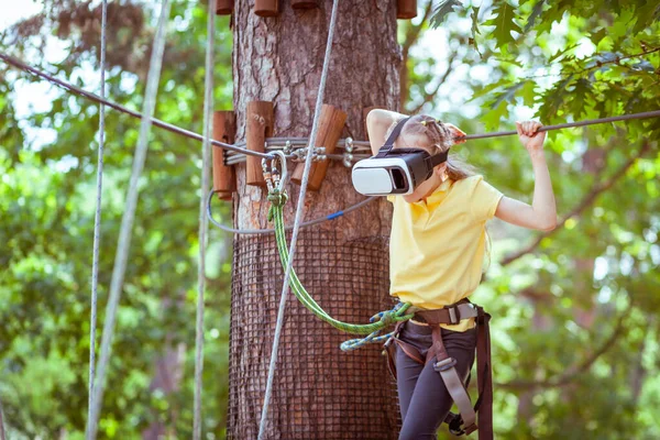 这个女孩玩一个虚拟游戏 她实际上是去参加体育活动 虚拟现实眼镜有助于体育运动 虚拟眼镜沉浸在孩子们的森林绳索探险公园里 — 图库照片