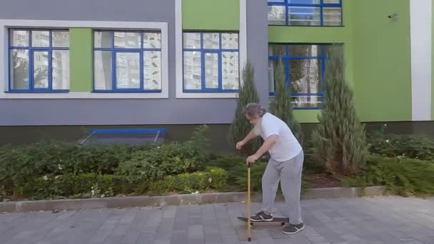 老爷爷在街上骑滑板 那位老人用手杖帮助他骑滑板 一位老人正在学习骑滑板 — 图库视频影像