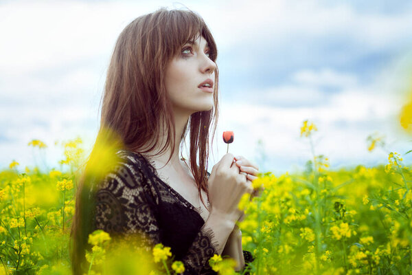 A young beautiful woman stands in a field with yellow flowers and is sad. Sad face of a beautiful woman.