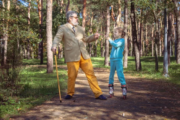 祖父は公園で孫娘と散歩する 女の子のローラーは 古い祖父と公園でスケートします 面白いおじいちゃんと小さな女の子が遊んで冗談を言っている ロイヤリティフリーのストック画像