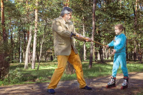祖父は公園で孫娘と散歩する 女の子のローラーは 古い祖父と公園でスケートします 面白いおじいちゃんと小さな女の子が遊んで冗談を言っている ロイヤリティフリーのストック写真